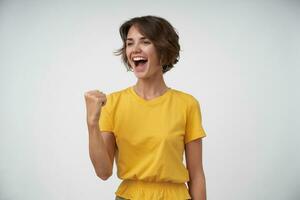 Overjoyed young brunette female with short haircut looking aside and smiling with wide mouth opened, raising fists happily in yes gesture, isolated over white background photo