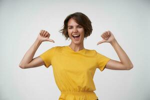 Portrait of self-confident lovely lady with casual hairstyle looking at camera joyfully and pointing happily to herself, standing over white background with raised hands photo