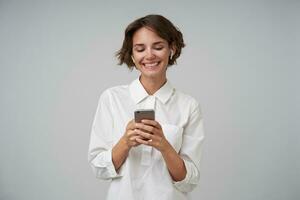 Cheerful young brunette lady with short haircut holding mobile phone in raised hands and looking happily at screen, wearing formal clothes while standing over white background photo