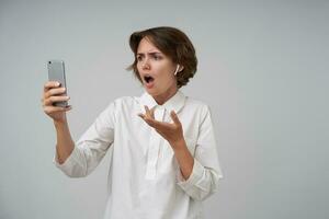 Portrait of young glum female with casual hairstyle holding mobile phone in raised hand and having video call, looking at screen with shocked face, posing over white background in formal clothes photo