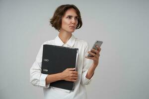 Thoughtful young brunette woman with casual hairstyle keeping mobile phone in hand and looking aside thoughtfully, making appointment on working day, posing over white office wall photo