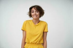 Beautiful young brunette woman with short haircut feeling shy and biting her underlip while looking at camera, wearing yellow t-shirt while posing over white background with hands down photo