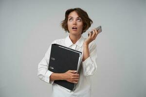 Indoor shot of dislpeased young brunette woman wearing formal clothes posing over white background with smartphone and folder with documents, having boring conversation and rolling eyes photo