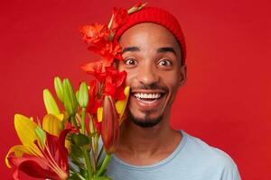 cerca arriba de joven alegre atractivo chico en rojo sombrero y azul camiseta, sostiene un ramo de flores en su manos, mira a el cámara con contento expresión y en general sonriente, soportes terminado rojo fondo foto