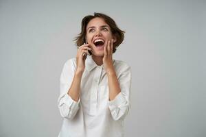 Overjoyed attractive young woman with short brown hair talking on phone and receiving good news, raising happily her palm to face, standing over white background in formal clothes photo