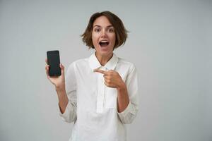 Joyful young attractive woman with short brown hair looking at screen positively with wide eyes opened, holding smartphone in hand and showing on it with forefinger, isolated over white background photo