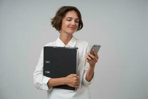 Portrait of pretty brunette woman with short haircut holding folder with documents and typing message on her mobile phone, being in nice mood, isolated over white background photo