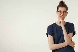 Portrait of young woman in t-shirt, keeps fist under chin and looks up aside, ponders the idea, consider different variants, thinking what do to solve the problem, troubled, over white background photo