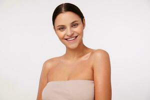 Indoor shot of cheerful young pretty brunette woman with natural makeup looking at camera with charming smile while standing against white background, being in nice mood photo