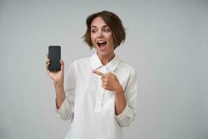 Cheerful young brunette woman with short haircut holding mobile phone in raised hand and pointing to it with excited face, wearing formal clothes while posing over white background photo