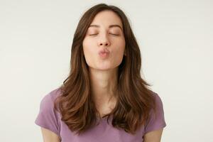 Sweet kiss directly to the camera. Portrait of pretty girl with blowing air kiss with pout lipsand closed eyes isolated on white background, shows tender feelings photo