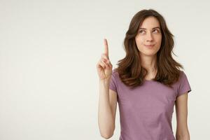 Portrait of attractive young woman looks to the left up corner, distance, smiles, ponders a thought or idea, thoughtful, keeps index finger up, wears casual t-shirt, on a white background photo