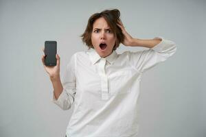 Shocked young brunette lady with casual hairstyle clutching her head and looking at camera surprisedly, holding mobile phone in raised hand while posing over white background photo