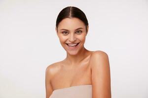 Happy young attractive dark haired green-eyed lady with ponytail hairstyle wearing nude top while posing over white background, looking cheerfully at camera with broad smile photo