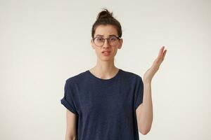 Studio shot of displeased student in glasses wears casual t-shirt looking camera feeling irritated with stupid nonsense standing unaware and clueless with one hand up over white background photo