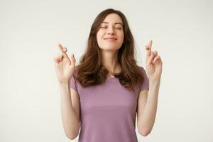 Portrait of young pretty inspired woman making a wish with crossed fingers, closed eyes, hopeful gesture, isolated on white studio background photo