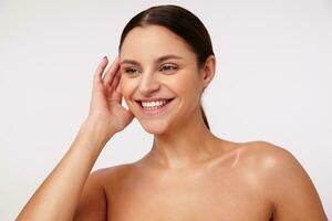 Cheerful lovely young brunette woman with ponytail hairstyle keeping raised hand behind her ear and smiling happily while posing against white background photo