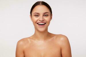 Cheerful attractive young dark haired lady with natural makeup wearing ponytail hairstyle while standing against white background, laughing happily while looking at camera photo