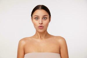 Surprised young pretty dark haired green-eyed woman with natural makeup looking amazedly at camera and raising eyebrows, posing over white background with naked shoulders photo