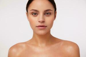 Close-up of beautiful young brunette green-eyed woman with natural makeup looking sincerely at camera, standing over white background with bare shoulders photo