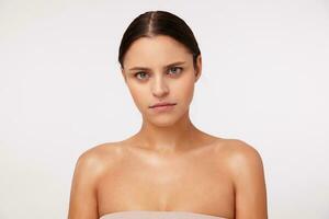 Close-up of gloomy young dark haired green-eyed lady with casual hairstyle looking seriously at camera and keeping lips folded, posing against white background with naked shoulders photo