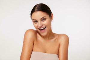 Charming young dark haired green-eyed woman with natural makeup looking happily at camera and smiling widely, dressed in beige top with opened shouders over white background photo