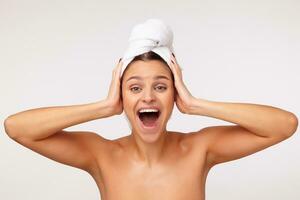 Excited young pretty lady with dark hair wrapped in bath towel clutching her head with raised hands and looking at camera with wide mouth opened, isolated over white background photo