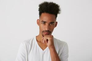 Pensive young attractive dark skinned unshaved guy keeping raised hand on his chin and looking thoughtfully at camera, isolated over white background photo