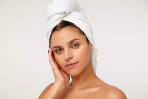 Closeup of charming young green-eyed dark haired female keeping raised palm on her cheek and looking gently at camera with light smile, posing after shower over white background photo