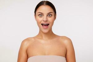 Portrait of surprised young cute brunette woman with casual hairstyle rounding eyes while looking at camera and keeping mouth wide opened, isolated over white background photo