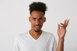 Indoor photo of young displeased dark skinned brunette guy with beard raising emotionally hand and grimacing face while standing over white background