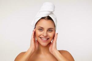 Closeup of attractive young lady with dark hair wrapped in bath towel touching her face with raised hands and looking cheerfully with broad smile, standing against white background photo