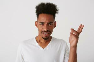 Studio shot of young pretty dark skinned unshaved curly guy frowning his face and keeping hand raised while looking discontentedly at camera, isolated over white background photo