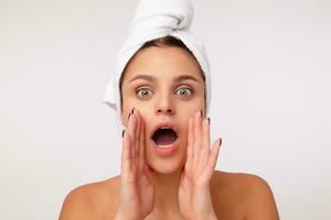 Indoor photo of lovely young dark haired female posing over white background with bath towel on her head, looking at camera with wide eyes opened and shouting something