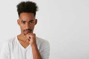 Portrait of young pretty brunette bearded man with dark skin holding his chin with raised hand while looking pensively at camera, posing over white background photo