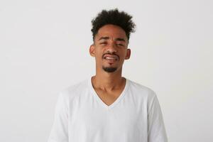 Studio shot of young handsome dark skinned bearded man with short curly hair squinting his eyes while looking languidly at camera, posing over white background photo