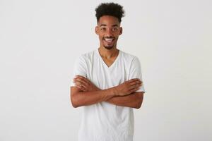 Portrait of young lovely dark skinned brunette guy dressed in basic t-shirt keeping hands crossed on his chest and smiling happily at camera, posing over white background photo