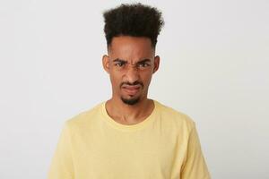 Portrait of displeased young brown-eyed brunette guy with dark skin frowning his face while looking discontentedly at camera, posing over white background photo