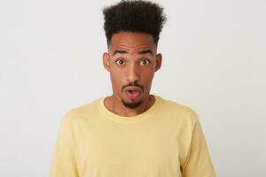 Portrait of dazed young attractive curly dark skinned unshaved guy rounding amazedly his brown eyes while looking amazedly at camera, isolated over white background photo