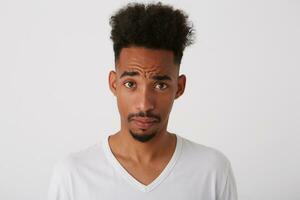 Portrait of bewildered young pretty unshaved man with dark skin frowning his forehead while looking confusedly at camera, isolated over white background photo