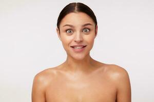 Open-eyed lovely young brunette woman with casual hairstyle looking amazedly at camera and raising surprisedly eyebrows while posing over white background with naked shoulders photo