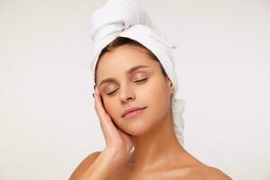 Pleasant looking young attractive brunette woman without makeup touching gently her face with raised hand and keeping eyes closed while standing over white background with towel on head photo
