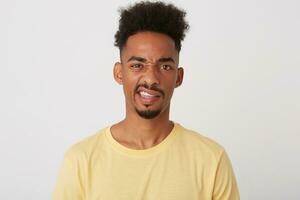 Studio shot of young pretty dark skinned unshaved guy with short trendy haircut making faces while standing over white background in yellow basic t-shirt photo