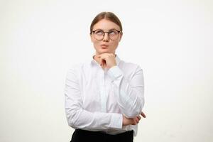 Pensive young lovely blonde female with ponytail hairstyle wearing glasses and formal clothes while standing over white background, leaning chin on raised hand while looking thoughtfully upwards photo