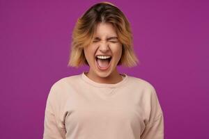 Indoor portrait of young dyed blonde female, with tattooed arms screaming with angry facial expression, isolated over purple background photo