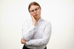 Upset young blonde woman with natural makeup frowning eyebrows and pouting lips while looking sadly at camera, keeping raised palm on her cheek while posing over white background photo