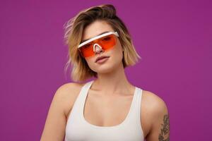 Indoor portrait of young dyed blonde female, with tattooed arms wears orange modern glasses, looks directly into camera with calm, emotionless facial expression, isolated over purple background photo