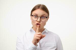 Excited young blonde woman in glasses raising hand with hush gesture while looking emotionally at camera, asking to keep silence while posing over white background photo