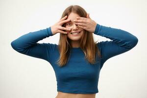 Portrait of young cheerful blonde female with natural makeup looking gladly at camera with broad smile and raising hands to her eyes, isolated over white background photo