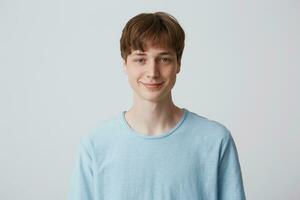 retrato de atractivo sonriente de ojos azules joven chico con corto Corte de pelo usa azul camiseta en pie y siente confidente aislado terminado blanco antecedentes mira directamente en cámara foto